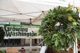 At the Phinney Center Farmers Market Holiday Market, a Growing Washington vendor sign hangs below a white tent, while a wreath made of greenery hangs in the foreground.