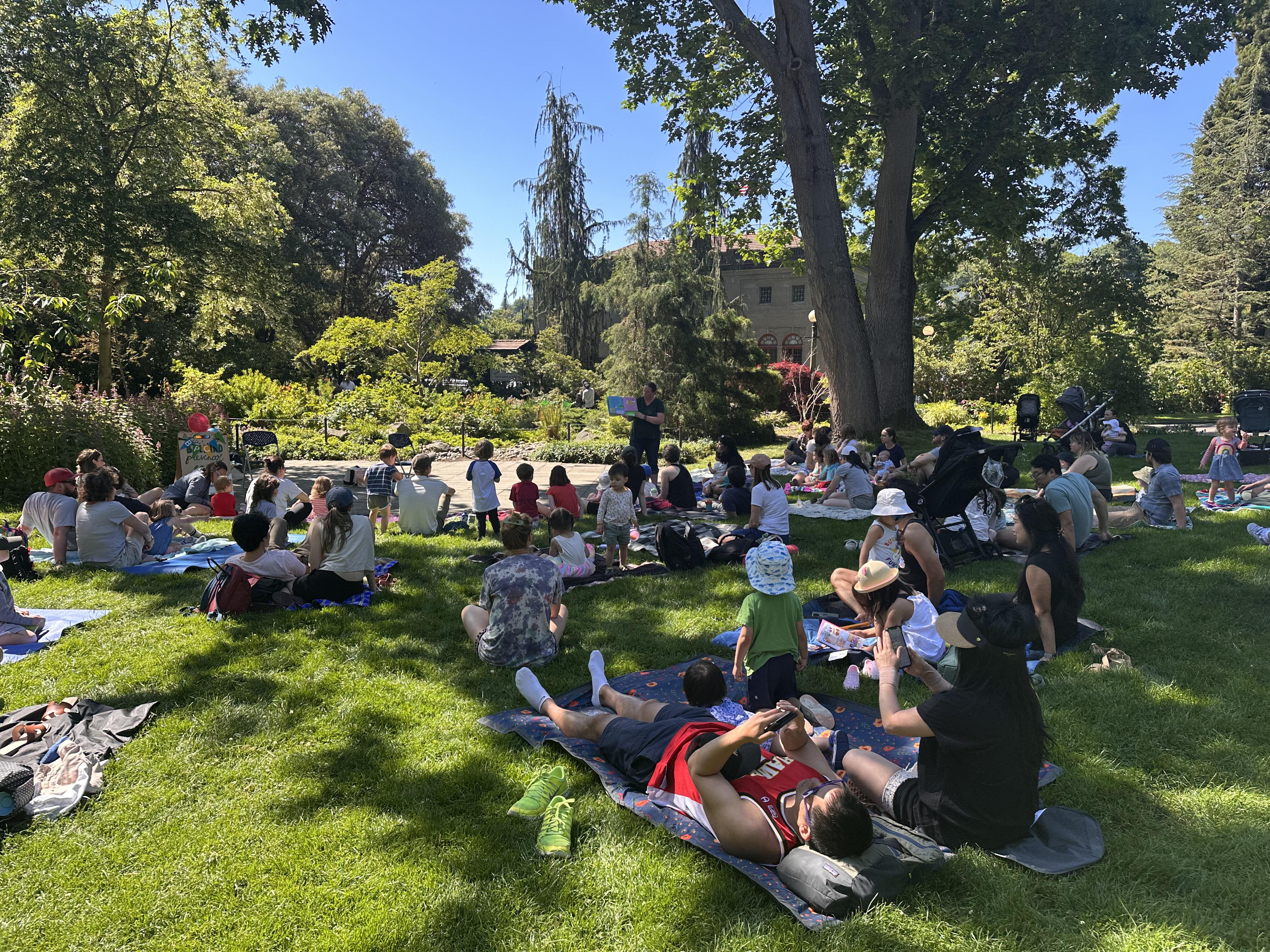 Families with young children sit on the grass at the Ballard Locks, under a big tree and blue sky, while an adult stands and reads from a book.